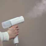 A close-up of a person using a handheld clothes steamer against a neutral background.